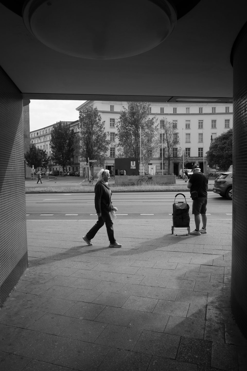 A pedestrian crossing in front of two people at a shaded bus stop
